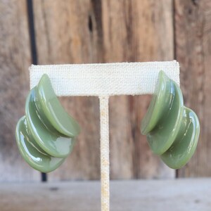 May include: A pair of olive green clip-on earrings. Each earring features a layered, curved design, creating a three-dimensional effect. The earrings have a glossy finish and are displayed on a neutral-colored stand against a wooden background.
