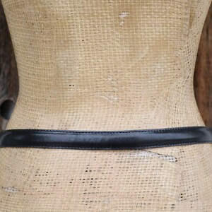 May include: A black leather belt is wrapped around a mannequin. The belt is thin and appears to be made of smooth leather. The mannequin is a light tan color and has a woven texture.