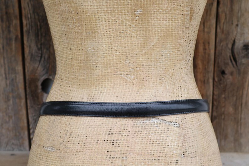 May include: A black leather belt is wrapped around a mannequin. The belt is thin and appears to be made of smooth leather. The mannequin is a light tan color and has a woven texture.