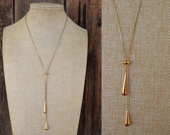 May include: A gold-toned necklace with a long, slender chain and a decorative pendant. The pendant features two teardrop-shaped elements suspended from a small, round bead. The necklace is displayed on a beige mannequin.