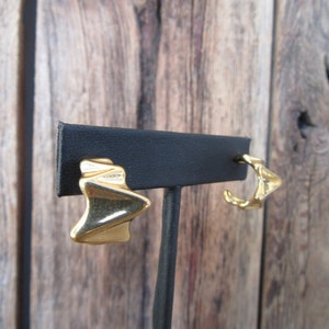 May include: A pair of gold-tone earrings with a geometric design. The earrings are displayed on a black stand.