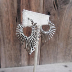 May include: A pair of silver hoop earrings with a sunburst design. The earrings are on a white display stand.