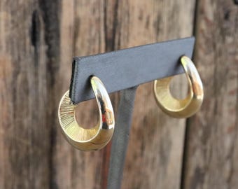 May include: Gold-toned hoop earrings with a teardrop shape. The earrings have a polished, reflective surface and a textured inner design. The earrings are displayed on a black earring stand against a wooden background.