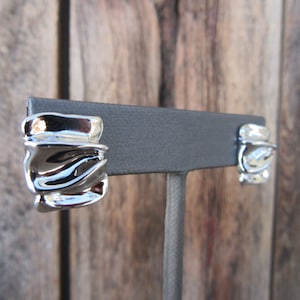 May include: A pair of silver tone earrings with a wavy, abstract design. The earrings are displayed on a black stand.