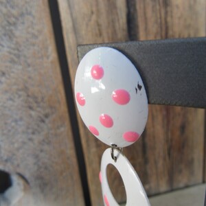 May include: White and pink polka dot earrings. The earrings are made of metal and have a shiny finish. The top earring is round and the bottom earring is a teardrop shape with a hole in the center.