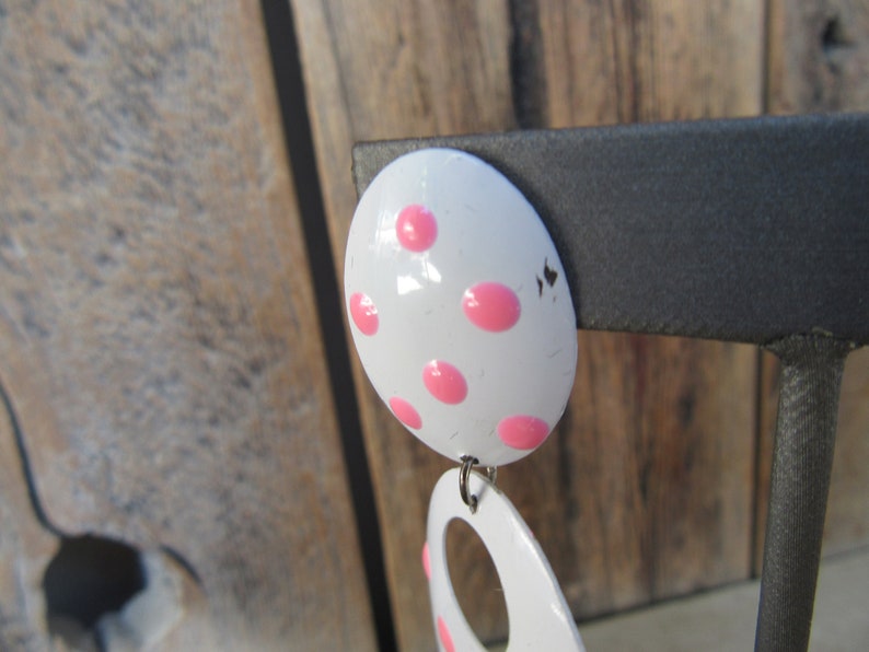 May include: White and pink polka dot earrings. The earrings are made of metal and have a shiny finish. The top earring is round and the bottom earring is a teardrop shape with a hole in the center.