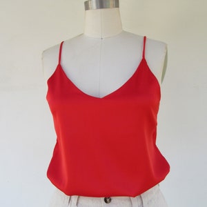 May include: A vibrant red camisole top with a V-neck design. The top is displayed on a mannequin, paired with light-colored linen pants. The thin straps and smooth fabric create a sleek, minimalist aesthetic. The top's bright color makes it a standout piece.