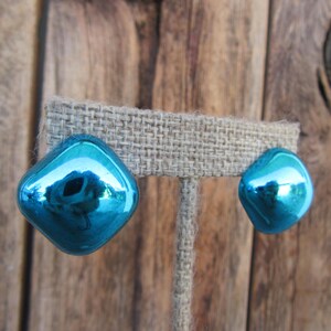 May include: A pair of teal blue, shiny, square-shaped earrings. The earrings are displayed on a burlap background.
