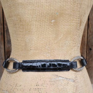 May include: A black belt with a glossy, crocodile-textured center section and ornate silver-toned circular buckles. The belt is displayed on a mannequin against a rustic wooden background.