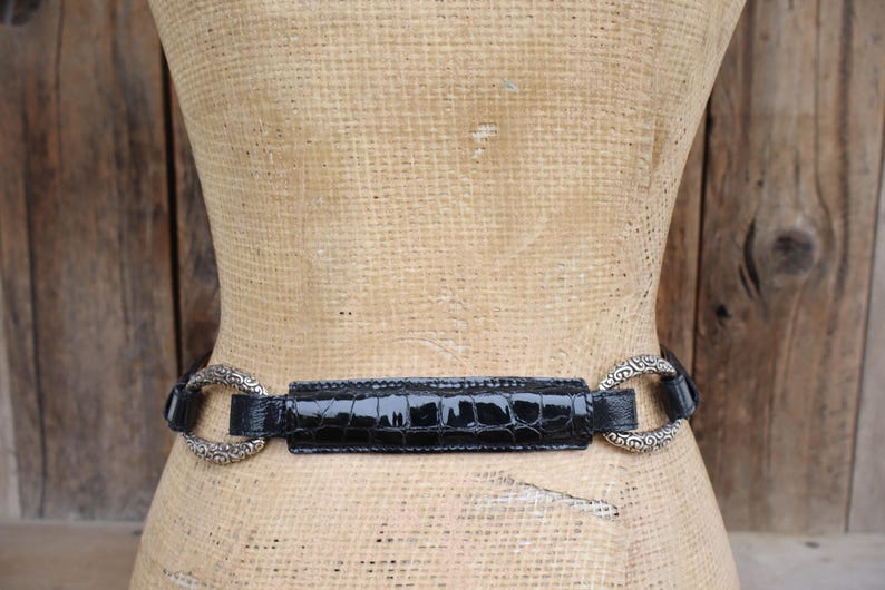 May include: A black belt with a glossy, crocodile-textured center section and ornate silver-toned circular buckles. The belt is displayed on a mannequin against a rustic wooden background.
