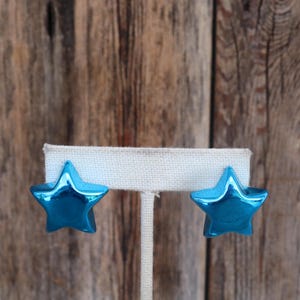 May include: A pair of blue star-shaped earrings on a white display stand. The earrings are made of a shiny metal and have a slightly 3D effect.