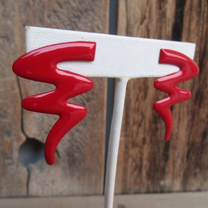 May include: A pair of red abstract shaped earrings on a white display stand. The earrings are made of a shiny, glossy material.