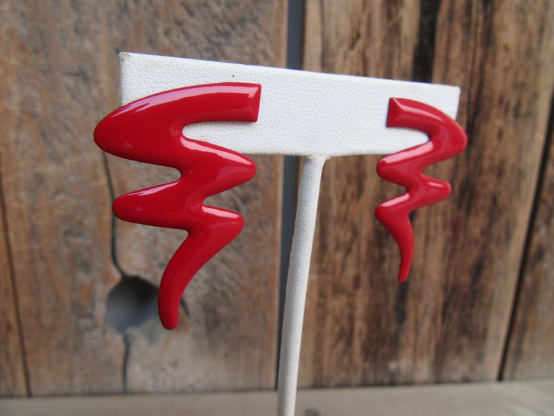 May include: A pair of red abstract shaped earrings on a white display stand. The earrings are made of a shiny, glossy material.