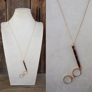 Puede incluir: Collar dorado con un colgante con forma de gafas. El colgante presenta una barra marrón con estampado de carey y dos círculos dorados. El collar se exhibe sobre un maniquí blanco.