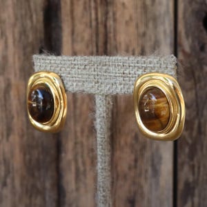K&ouml;nnte beinhalten: Ein Paar goldfarbene Clip-Ohrringe. Jeder Ohrring hat eine rechteckige Form mit abgerundeten Ecken, die einen braunen, oval geformten Stein einrahmt. Die Ohrringe werden auf einem neutralfarbenen Stoff pr&auml;sentiert.