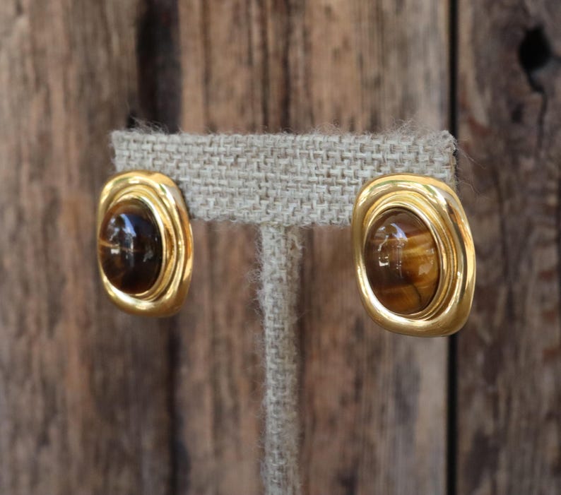 K&ouml;nnte beinhalten: Ein Paar goldfarbene Clip-Ohrringe. Jeder Ohrring hat eine rechteckige Form mit abgerundeten Ecken, die einen braunen, oval geformten Stein einrahmt. Die Ohrringe werden auf einem neutralfarbenen Stoff pr&auml;sentiert.