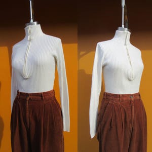 May include: A white ribbed turtleneck sweater with a zipper up the front. The sweater is worn with brown corduroy pants.