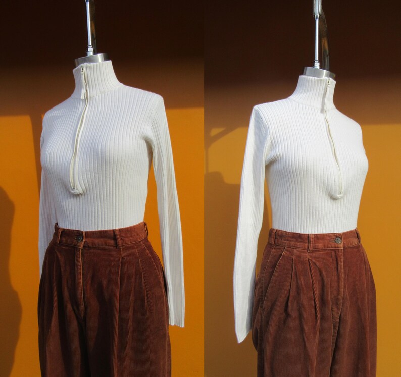 May include: A white ribbed turtleneck sweater with a zipper up the front. The sweater is worn with brown corduroy pants.