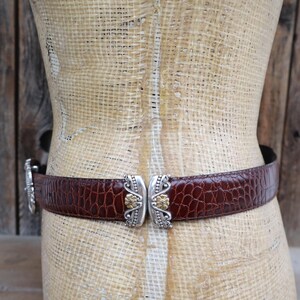 May include: A brown leather belt with a crocodile texture is fastened around a mannequin. The belt features a silver clasp with gold-colored accents. The background is a rustic wooden wall.