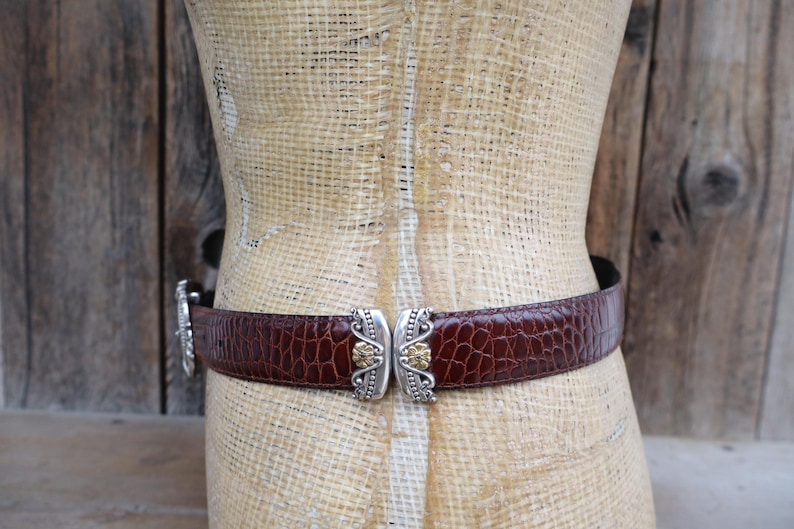 May include: A brown leather belt with a crocodile texture is fastened around a mannequin. The belt features a silver clasp with gold-colored accents. The background is a rustic wooden wall.