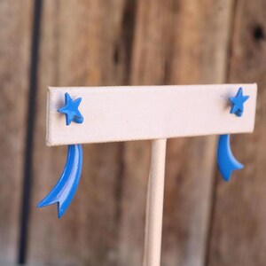 May include: A pair of bright blue earrings featuring a five-pointed star stud design with a curved, ribbon-like tail extending downward from each star. The earrings have a smooth, glossy finish and are displayed on a light-colored stand.