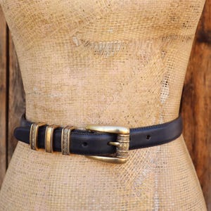 May include: A black leather belt with gold-toned metal accents. The belt features a gold buckle and decorative gold rings. The belt is displayed on a mannequin against a wooden background.
