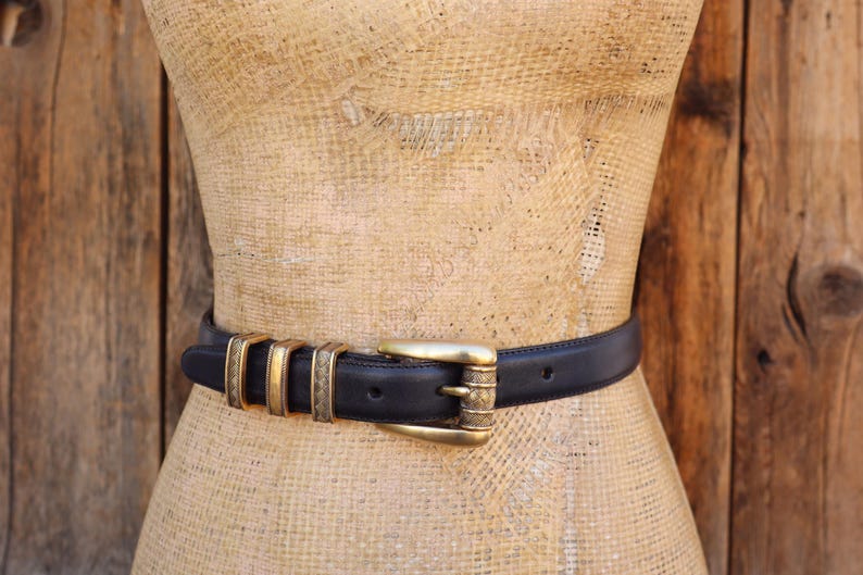 May include: A black leather belt with gold-toned metal accents. The belt features a gold buckle and decorative gold rings. The belt is displayed on a mannequin against a wooden background.