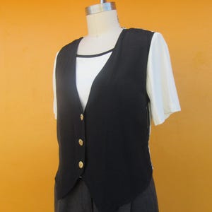 Peut inclure: Un gilet noir sans manches avec des boutons dor&eacute;s. Le gilet a un col en V et est port&eacute; sur une chemise blanche. Le gilet est associ&eacute; &agrave; un pantalon gris.