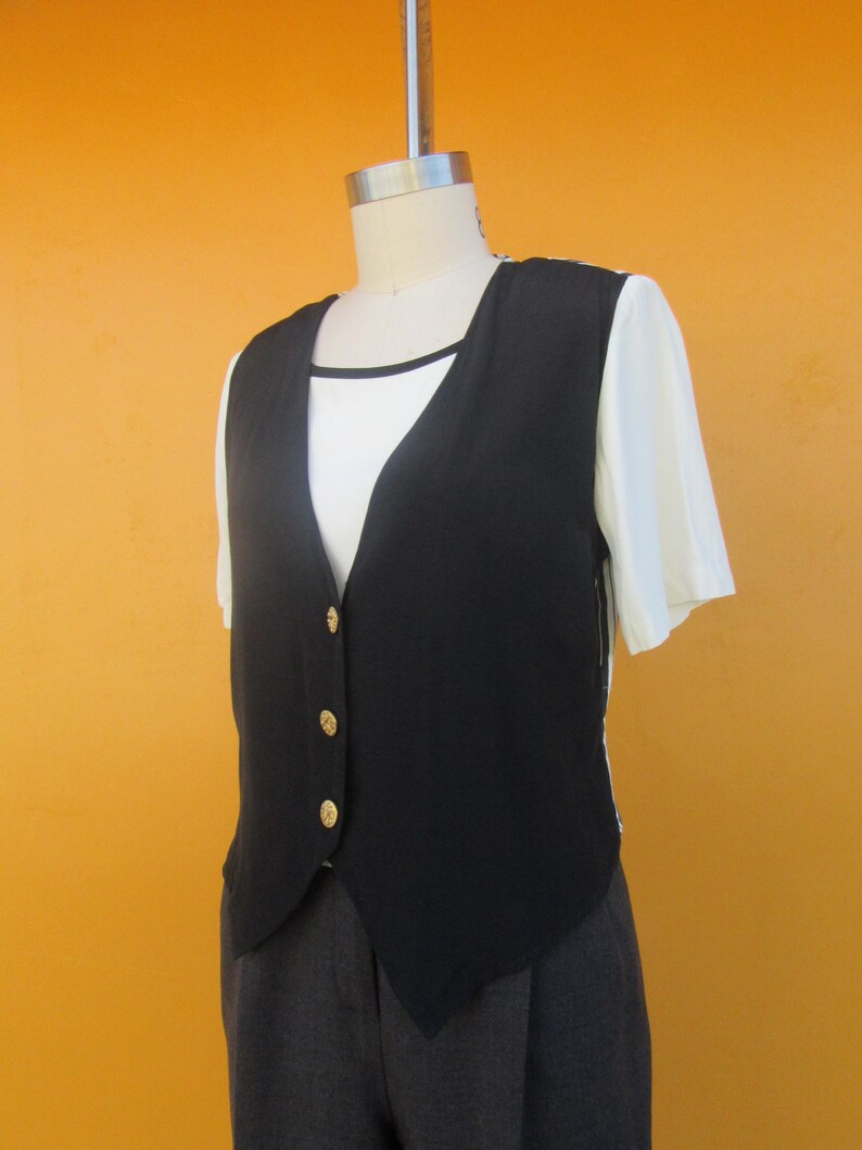 Peut inclure: Un gilet noir sans manches avec des boutons dor&eacute;s. Le gilet a un col en V et est port&eacute; sur une chemise blanche. Le gilet est associ&eacute; &agrave; un pantalon gris.