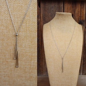 May include: A silver-toned necklace with a long, slender pendant featuring two geometric shapes. The necklace is displayed on a tan mannequin, set against a neutral backdrop. The chain is delicate and the pendant has a modern, minimalist design.