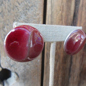 Op de afbeelding: Een paar rode keramische oorbellen met een glanzende afwerking. De oorbellen worden gepresenteerd op een witte standaard.