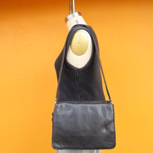 May include: A black leather shoulder bag with a rectangular silhouette and a fold-over flap closure. The bag features a long, thin shoulder strap and is displayed on a mannequin wearing a dark sleeveless knit top and light-colored trousers.