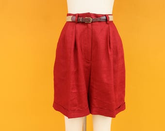 Puede incluir: Unos shorts rojos de cintura alta con cinturón a juego. Los shorts tienen la parte delantera plisada y dobladillos con vuelta. El cinturón es de cuero marrón con una hebilla dorada. Los shorts se exhiben en un maniquí sobre un fondo amarillo.