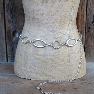 May include: Silver-toned chain belt with oval and circular links. The belt is draped on a mannequin, showcasing its design. A longer chain hangs from the belt, adding a decorative element. The background is a rustic wooden surface.