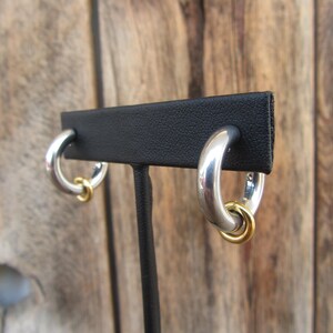 May include: A pair of silver hoop earrings with a gold accent ring. The earrings are displayed on a black stand.