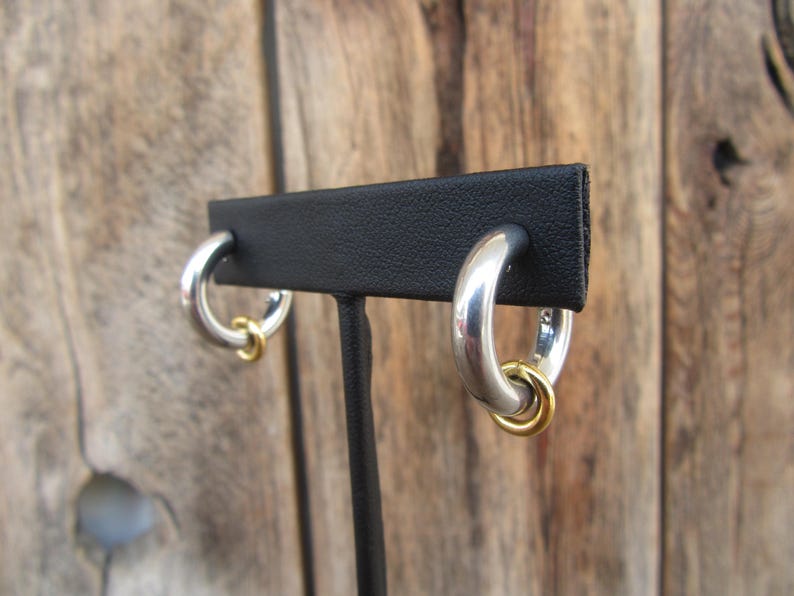 May include: A pair of silver hoop earrings with a gold accent ring. The earrings are displayed on a black stand.