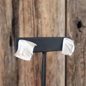 May include: A pair of silver-toned stud earrings with a curved, wing-like design. The earrings are displayed on a black earring stand against a rustic wooden background. The earrings have a smooth, polished finish.