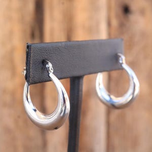 May include: A pair of polished silver hoop earrings. The earrings have a smooth, rounded design and are displayed on a black earring stand against a blurred wooden background. The earrings are a classic jewelry item.