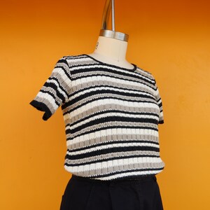May include: A striped short-sleeved shirt with black, white and beige stripes. The shirt is made of a soft, ribbed fabric and has a crew neckline.