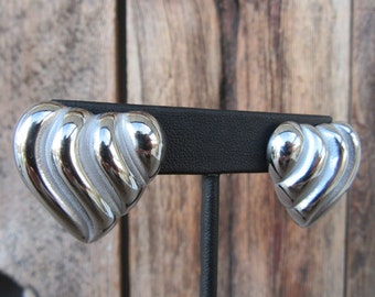 May include: A pair of silver heart-shaped earrings with a ridged design. The earrings are displayed on a black stand.