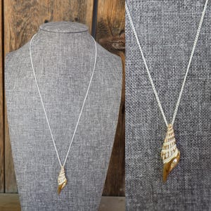 May include: A gold-toned seashell pendant necklace with a white chain. The pendant is a detailed replica of a seashell, with a pearl accent. The necklace is displayed on a gray mannequin, against a wooden background.