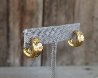 May include: A pair of gold-colored hoop earrings. The earrings are displayed on a gray fabric earring holder. The earrings have a smooth, curved design and a shiny finish. The background is a blurred wooden surface.