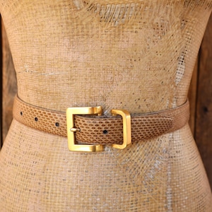 May include: A tan belt with a textured, reptilian-like pattern and a square, gold-toned metal buckle. The belt features a series of evenly spaced adjustment holes and is secured with a matching gold-toned metal keeper loop for a clean finish.
