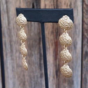 May include: Gold-toned, textured dangle earrings. Each earring features a series of four textured, oval-shaped elements suspended from a round top. The earrings are displayed on a black earring stand against a blurred wooden background.