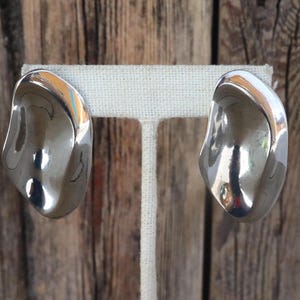 May include: A pair of polished silver-toned earrings. Each earring has a smooth, curved, organic shape, reflecting light. The earrings are displayed on a white fabric stand against a wooden background. These are statement earrings.