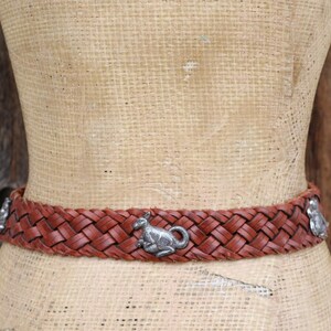 May include: A woven brown leather belt featuring silver kangaroo embellishments. The belt is displayed on a mannequin against a wooden backdrop. A fashionable accessory, approx. 3 cm wide.