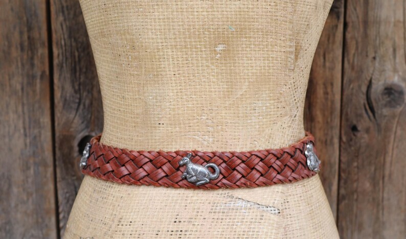 May include: A woven brown leather belt featuring silver kangaroo embellishments. The belt is displayed on a mannequin against a wooden backdrop. A fashionable accessory, approx. 3 cm wide.