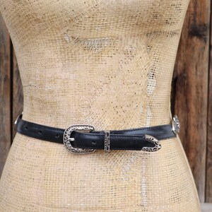 May include: A black leather belt with a silver-colored buckle and decorative accents. The belt is displayed on a mannequin against a rustic wooden background. The buckle and accents have an ornate, western-style design.