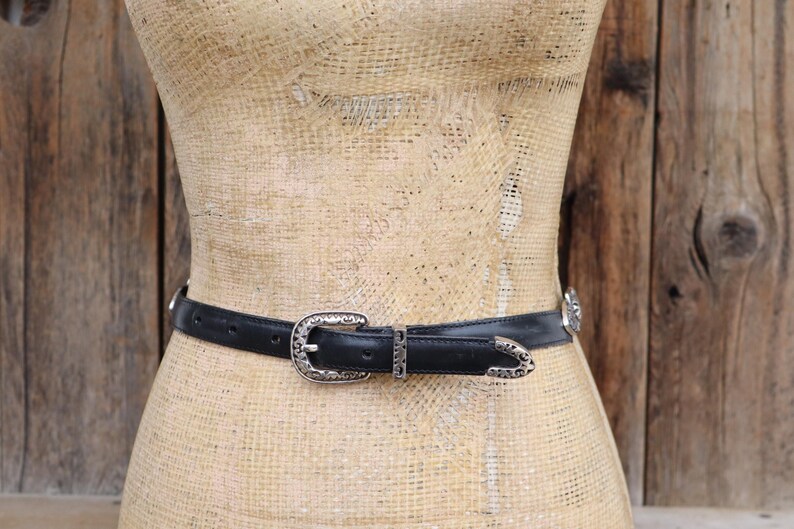 May include: A black leather belt with a silver-colored buckle and decorative accents. The belt is displayed on a mannequin against a rustic wooden background. The buckle and accents have an ornate, western-style design.