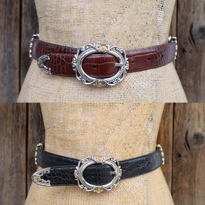 May include: Two belts are displayed, one brown and one black, each with a textured, crocodile-like pattern. Both belts feature an oval silver buckle with a decorative design and small gold-colored accents. The belts are displayed on a mannequin.